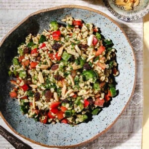 A bowl of colorful bulgur salad with diced vegetables, herbs, raisins, and slivered almonds sits on a patterned cloth next to a small dish of extra almonds and a glass of drink.