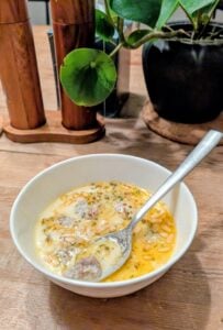 A white bowl filled with yogurt soup with pasta and meatballs, topped with herbs and melted cheese, sits on a wooden table. A spoon rests in the bowl. In the background, there are a potted plant and wooden salt and pepper grinders.