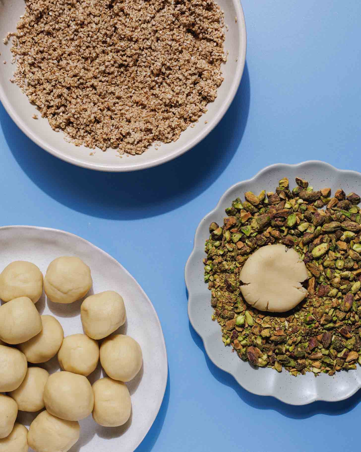 Three plates on a blue background: one with crumbled mixture, one with whole pistachios around a flat dough disc, and one with round dough balls ready for baking or decorating.