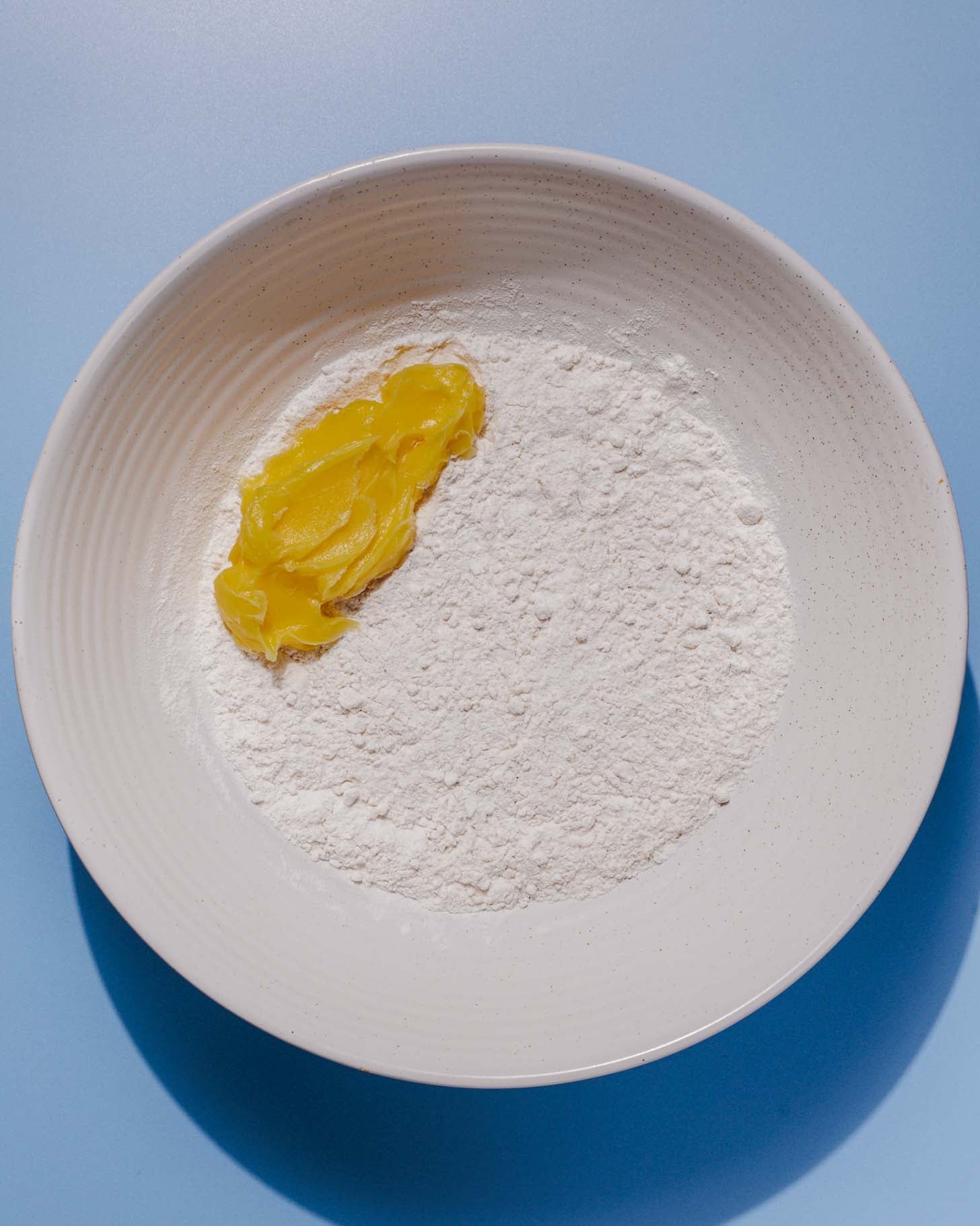 A white bowl containing a pile of white flour and a dollop of yellow ghee or butter, set on a light blue surface.