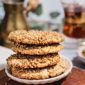 A stack of four sesame-coated cookies sits on a small white plate atop a wooden surface, with a blurred background featuring a glass of tea and a brass pot.