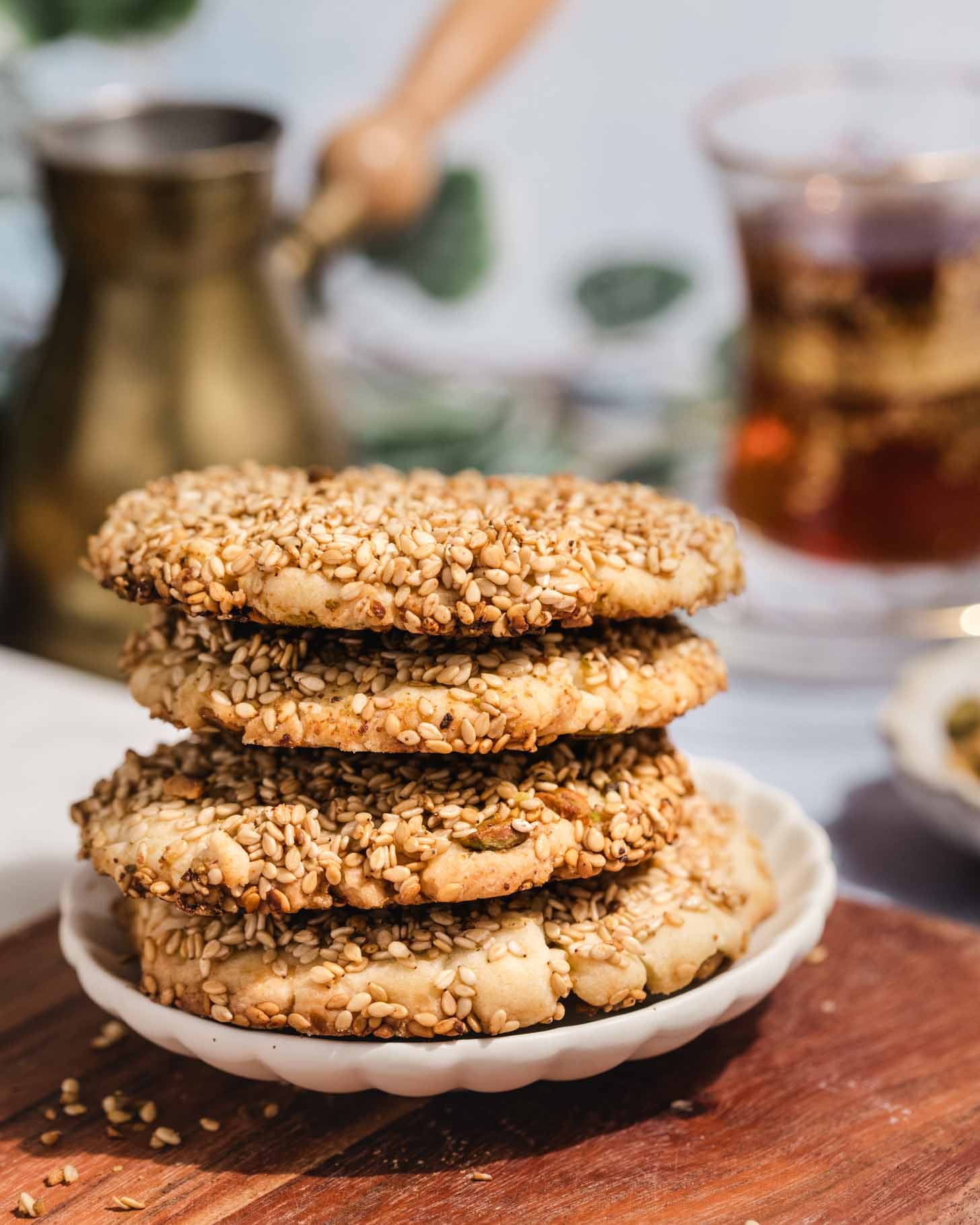 A stack of four sesame-coated cookies sits on a small white plate atop a wooden surface, with a blurred background featuring a glass of tea and a brass pot.