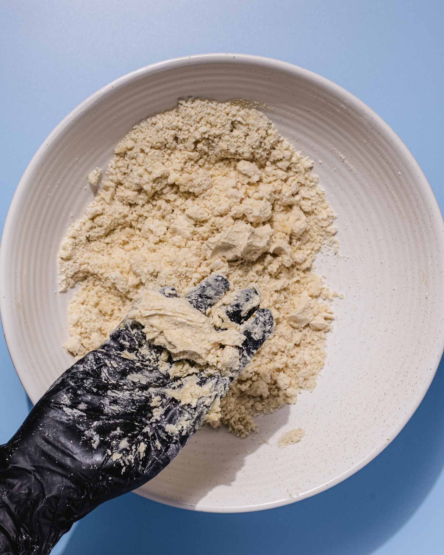 A hand wearing a black glove holds a crumbly mixture over a white bowl filled with more of the same mixture, set against a light blue background.