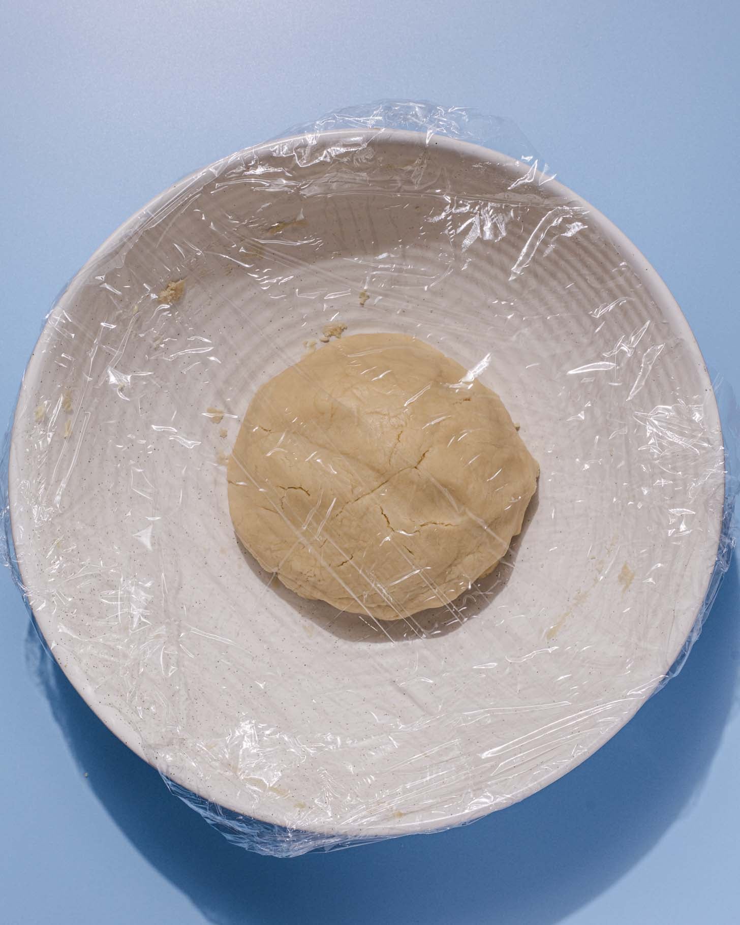 A ball of dough sits in a large white bowl covered with plastic wrap, placed on a light blue surface.