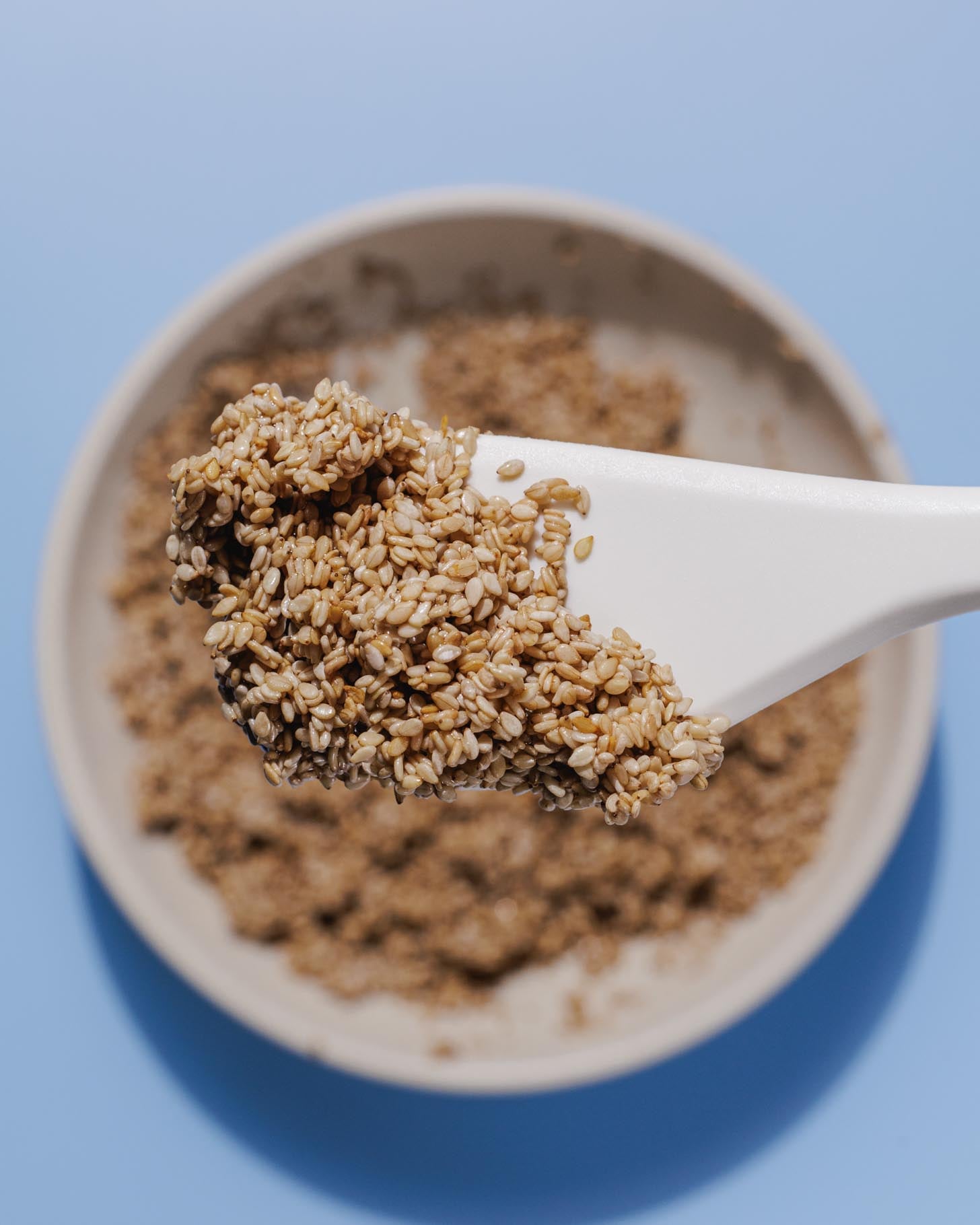 A white spoon holding a heap of sesame seeds above a bowl filled with more sesame seeds, set against a light blue background.