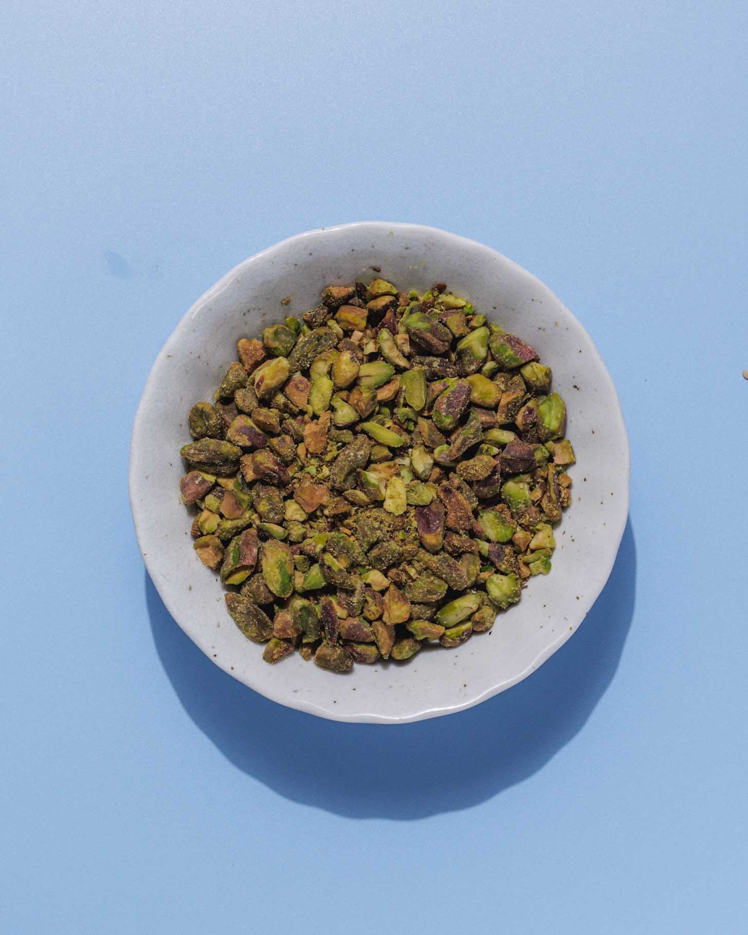 A small white bowl filled with chopped pistachios sits on a light blue surface. The pistachios are green and brown, and the bowl casts a faint shadow.