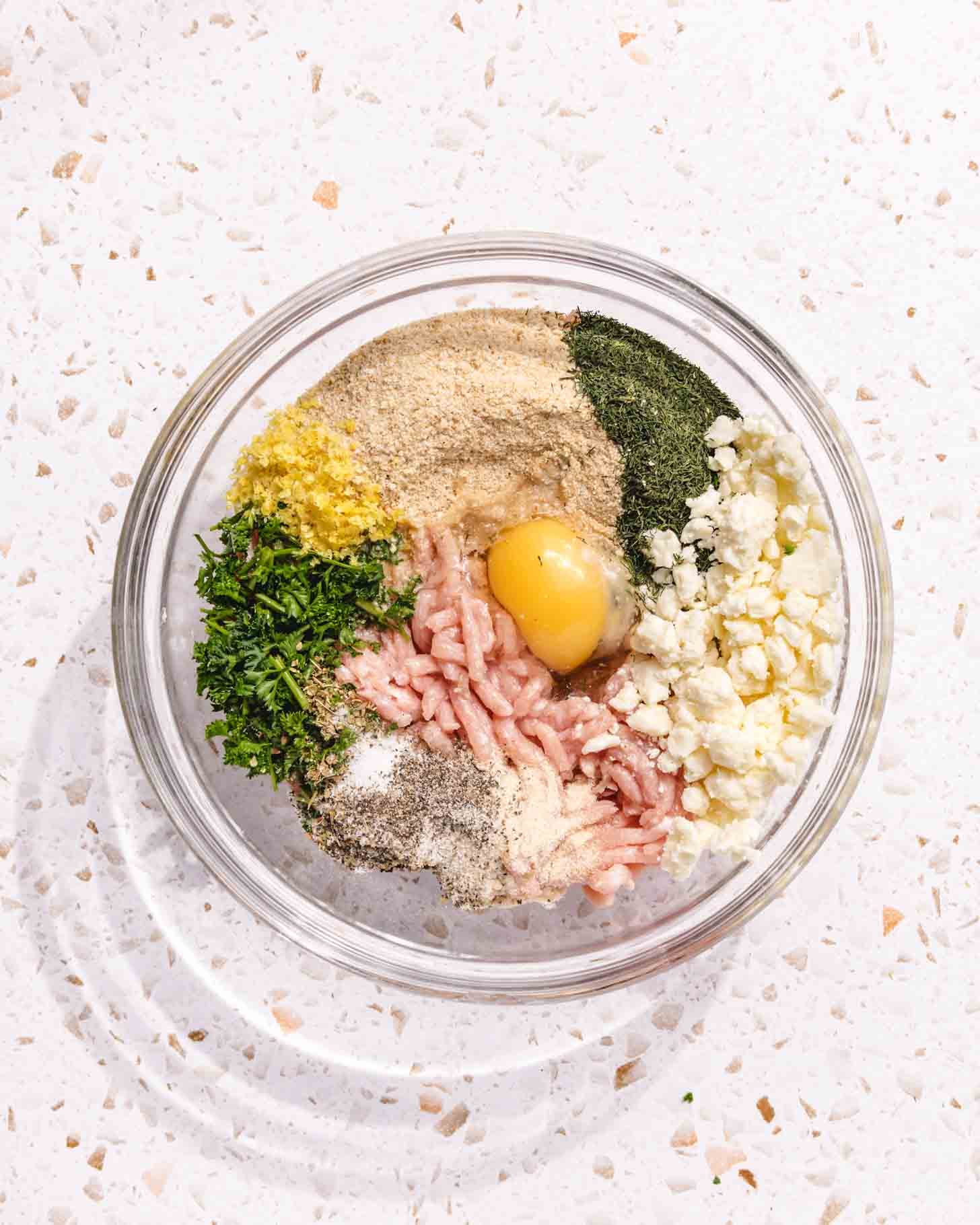 A glass bowl with ground meat, an egg, breadcrumbs, chopped herbs, crumbled cheese, and seasonings arranged in sections on a white speckled countertop.