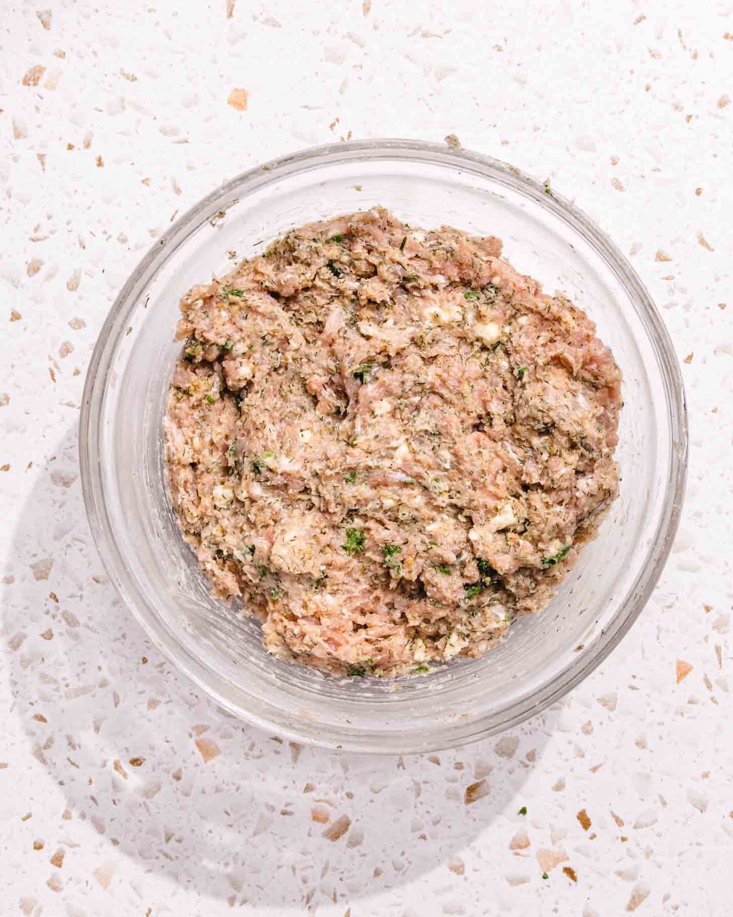 A clear glass bowl filled with a mixture of ground meat, herbs, and seasonings sits on a speckled white countertop.