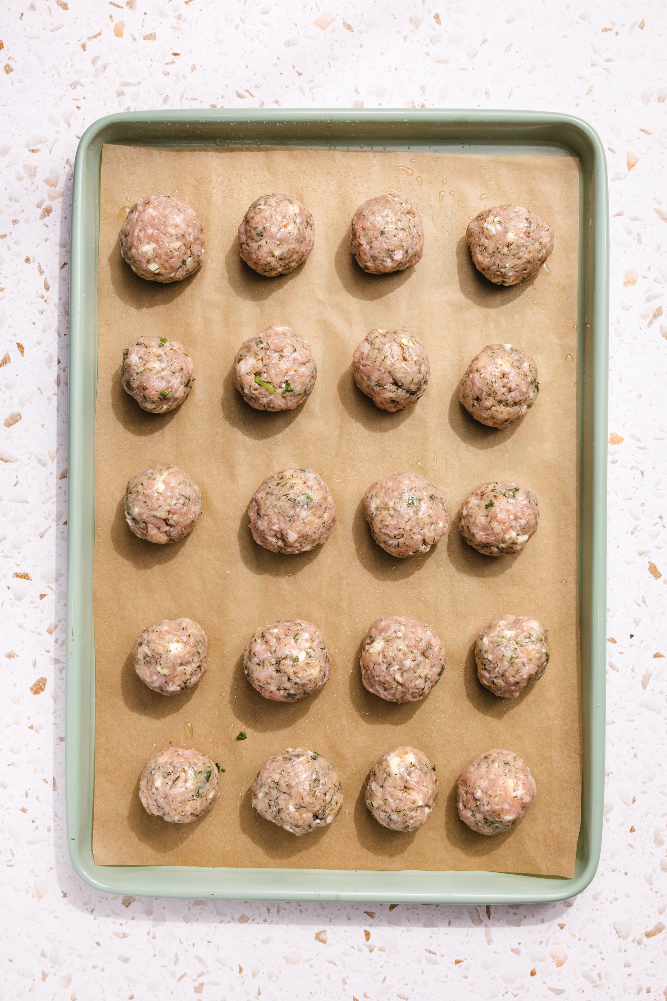 A green baking sheet lined with brown parchment paper holds 20 evenly spaced, uncooked meatballs made with herbs. The sheet rests on a white, speckled countertop.