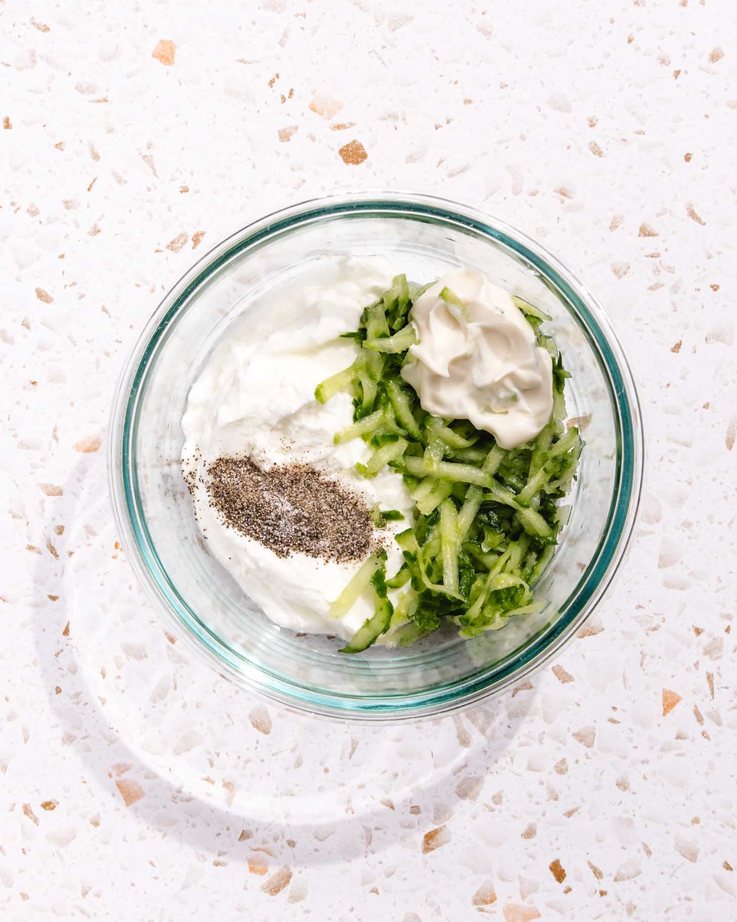 A glass bowl on a speckled countertop containing yogurt, shredded cucumber, mayonnaise, and ground black pepper, all not yet mixed together.