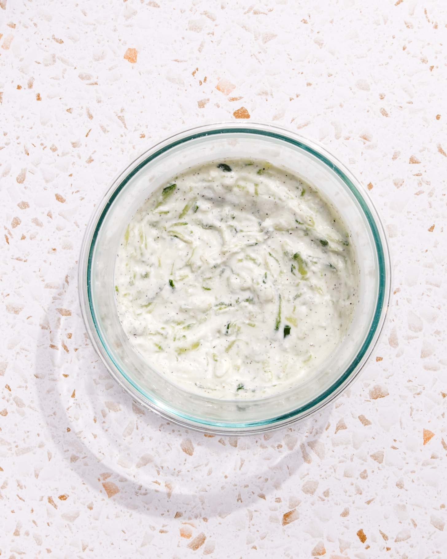 A glass bowl filled with creamy tzatziki sauce, showing visible shreds of cucumber and specks of herbs, sits on a light-colored speckled countertop.