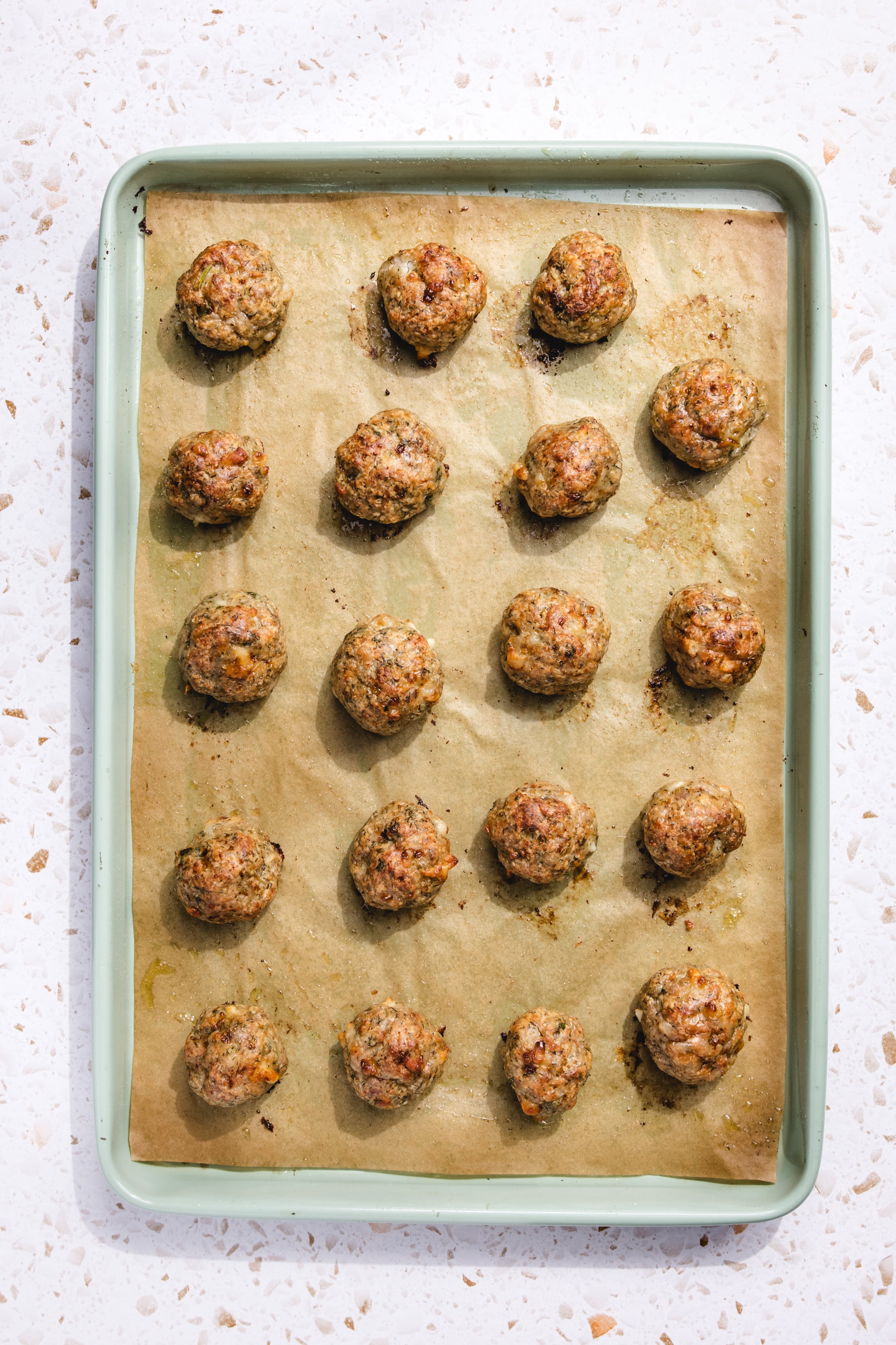 A green baking tray lined with brown parchment paper holds 20 evenly spaced, baked meatballs on a white speckled surface.