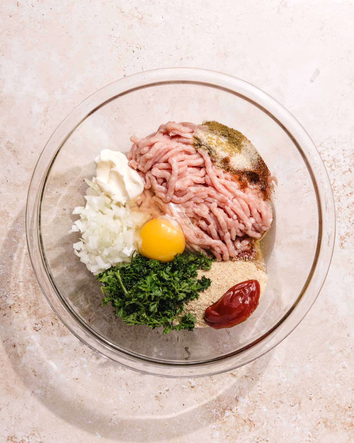A glass bowl containing ground meat, a raw egg, chopped onion, parsley, mayonnaise, ketchup, and various seasonings on a light countertop.