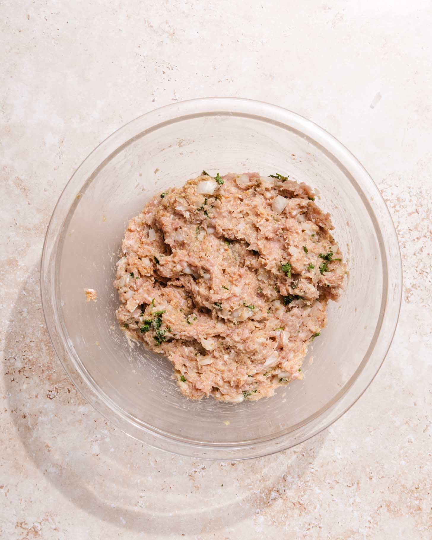 A clear glass bowl filled with a mixture of ground meat, chopped herbs, and onions sits on a light, textured surface.