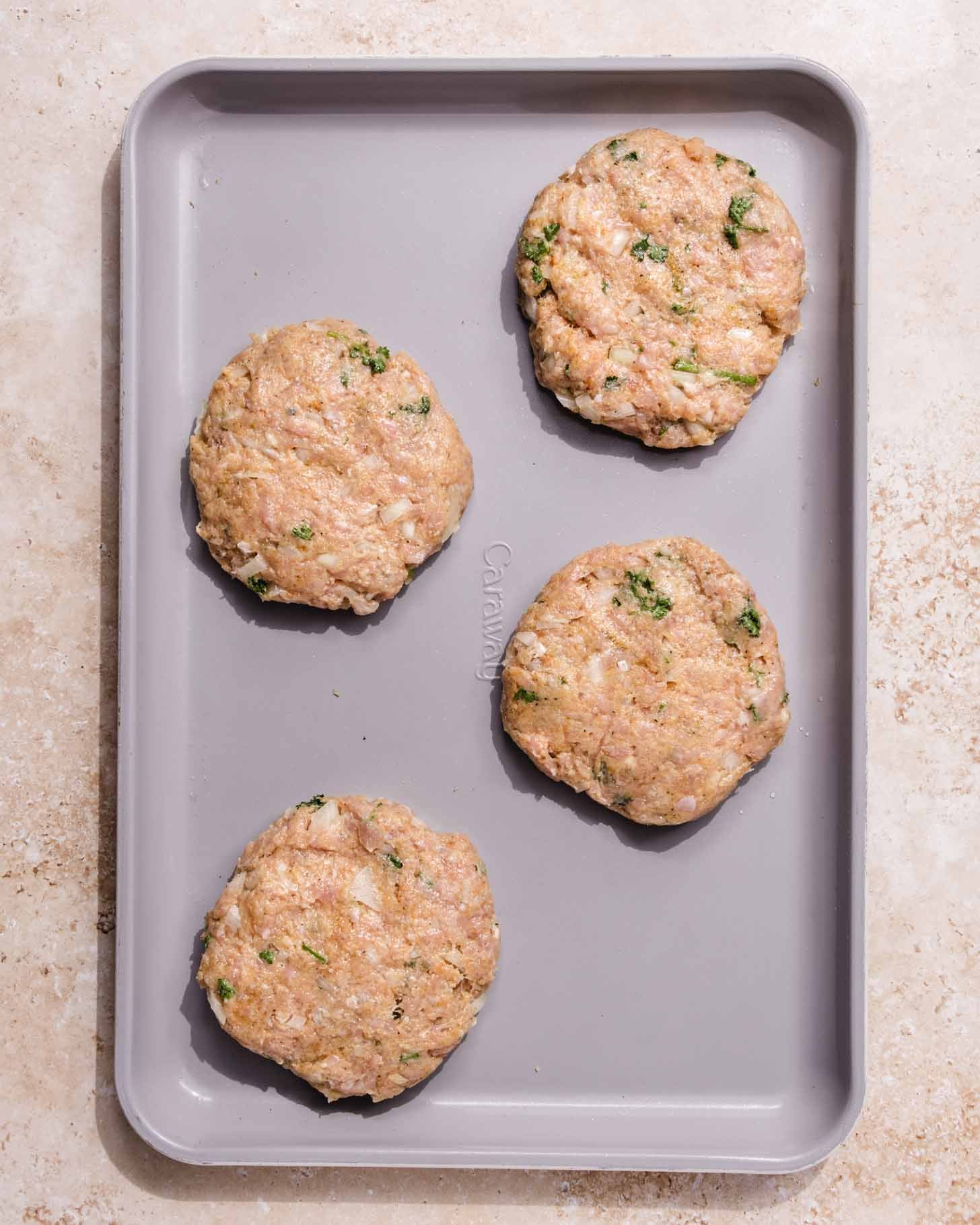 A gray baking sheet with four raw, round chicken patties mixed with herbs, placed evenly apart on a light-colored surface.