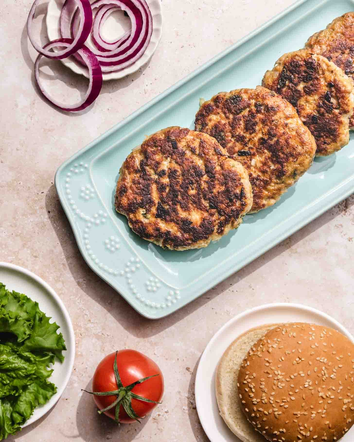 Three cooked burger patties on a light blue platter, with a sliced red onion, lettuce, a whole tomato, and a sesame seed hamburger bun beside them on a light countertop.