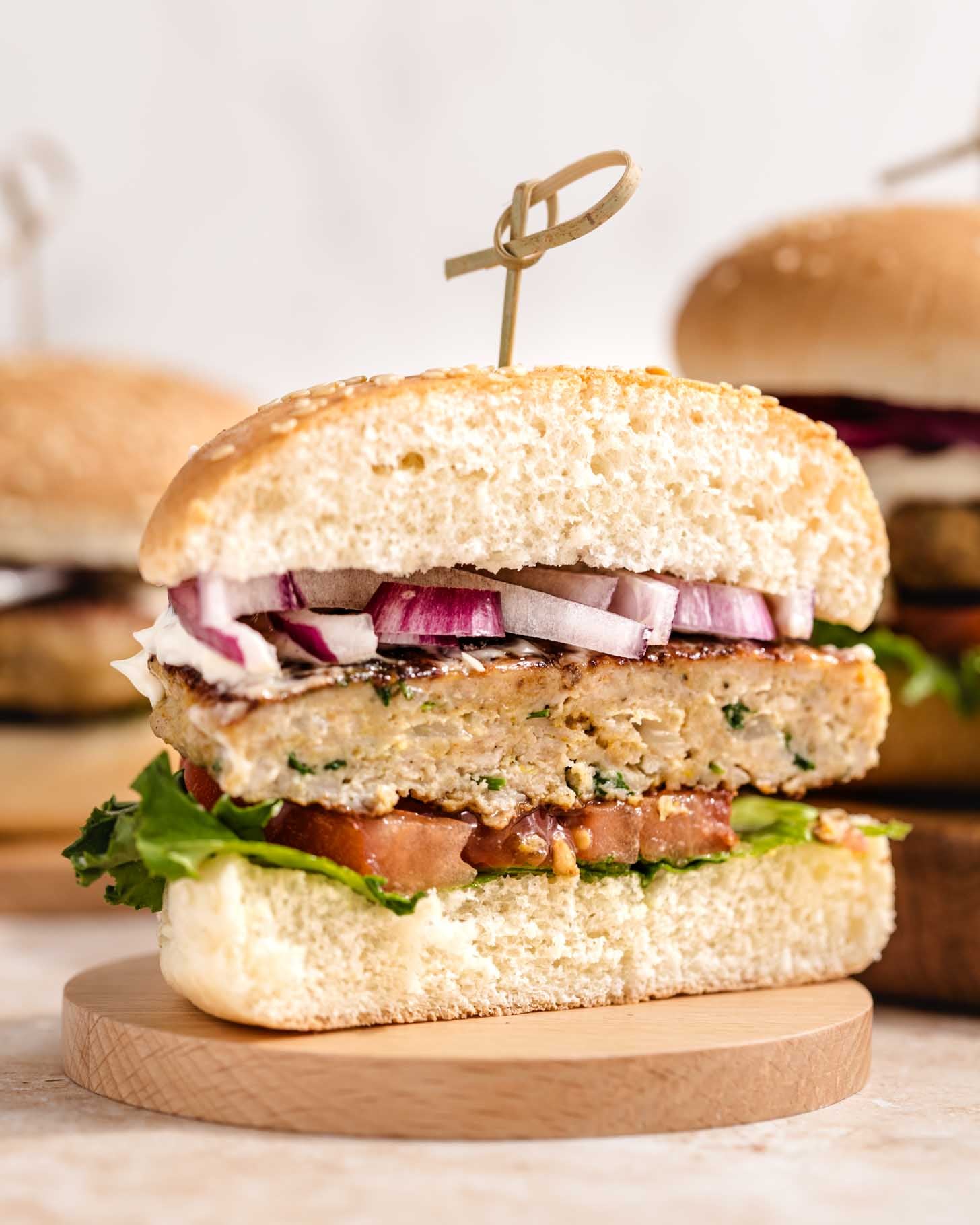 A sliced chicken burger on a wooden coaster, showing layers of lettuce, tomato, chicken patty, chopped red onion, and sauce inside a sesame seed bun, secured with a wooden pick.