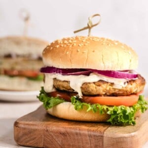 A grilled chicken burger with lettuce, tomato, onion, and creamy sauce on a sesame bun sits on a wooden board. Another burger is blurred in the background.