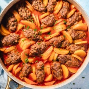 A skillet filled with oval-shaped meat patties and sliced potatoes cooked in a red tomato sauce, garnished with chopped herbs. The dish is on a light blue surface next to a plate of fresh herbs and a glass of water.