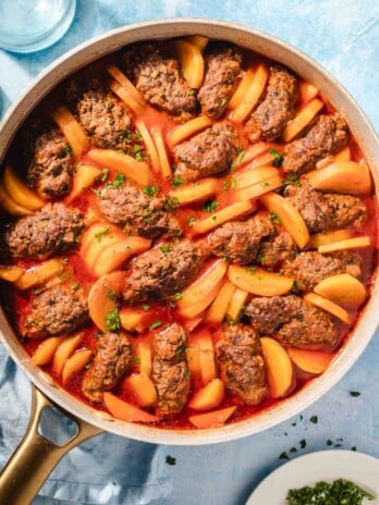 A skillet filled with oval-shaped meat patties and sliced potatoes cooked in a red tomato sauce, garnished with chopped herbs. The dish is on a light blue surface next to a plate of fresh herbs and a glass of water.