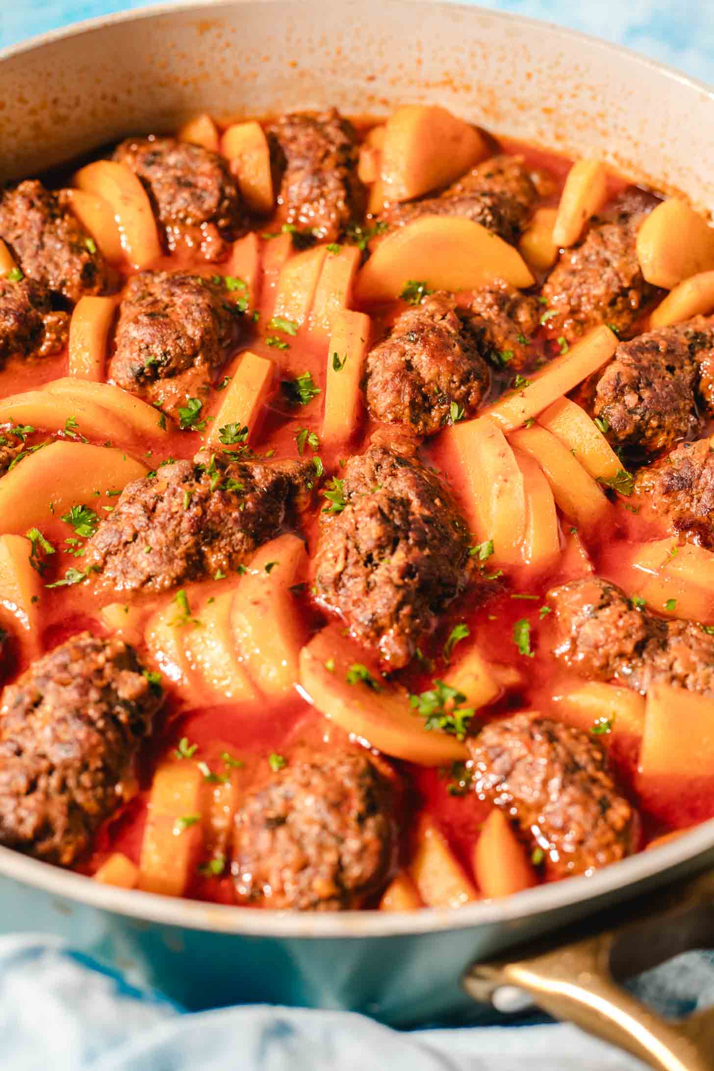 A pan filled with oval-shaped meatballs and sliced potatoes in a rich tomato sauce, garnished with chopped fresh herbs.