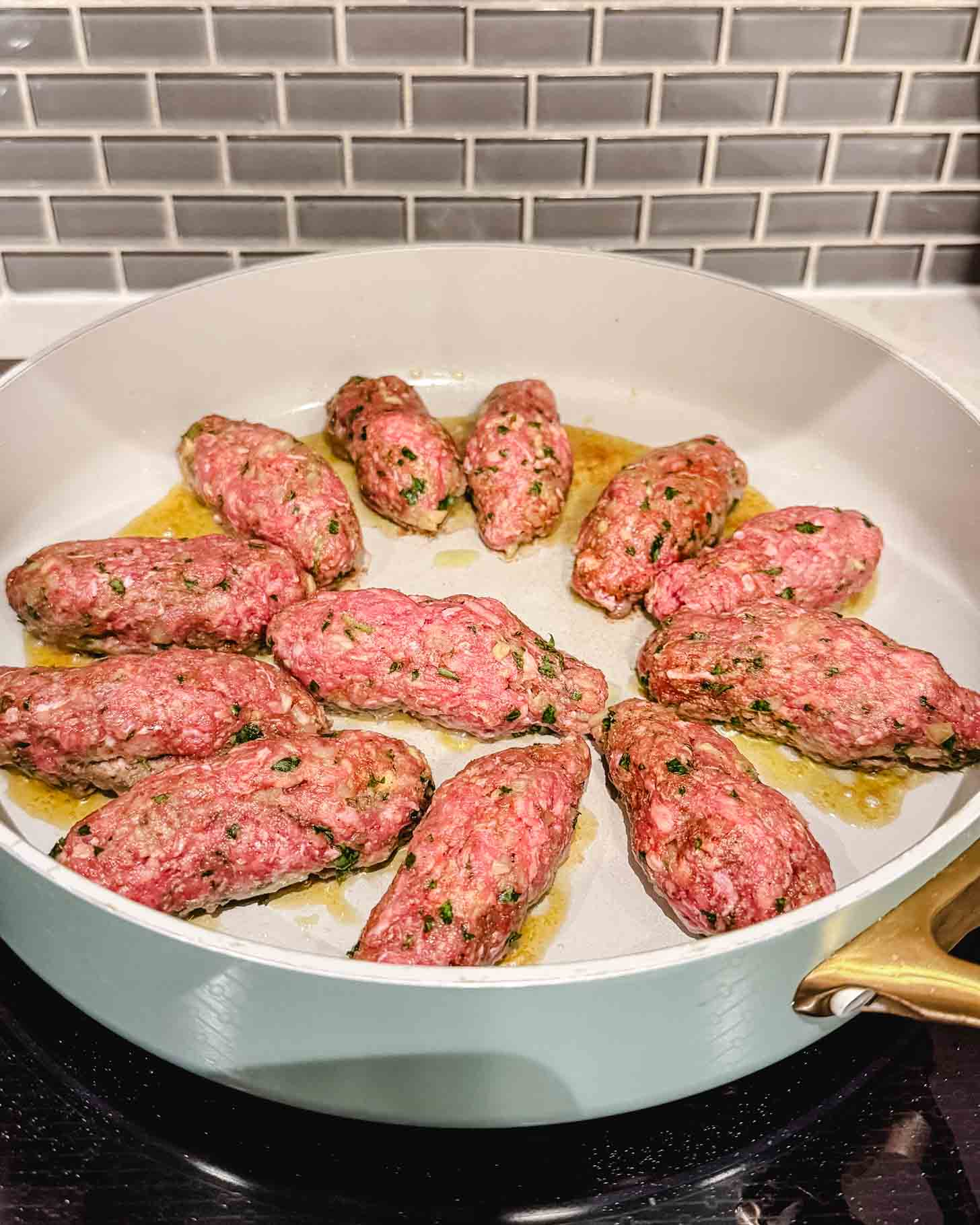 Raw, seasoned ground meat shaped into logs is cooking in a shallow white pan with some oil. The pan sits on a stovetop in front of a gray subway tile backsplash.
