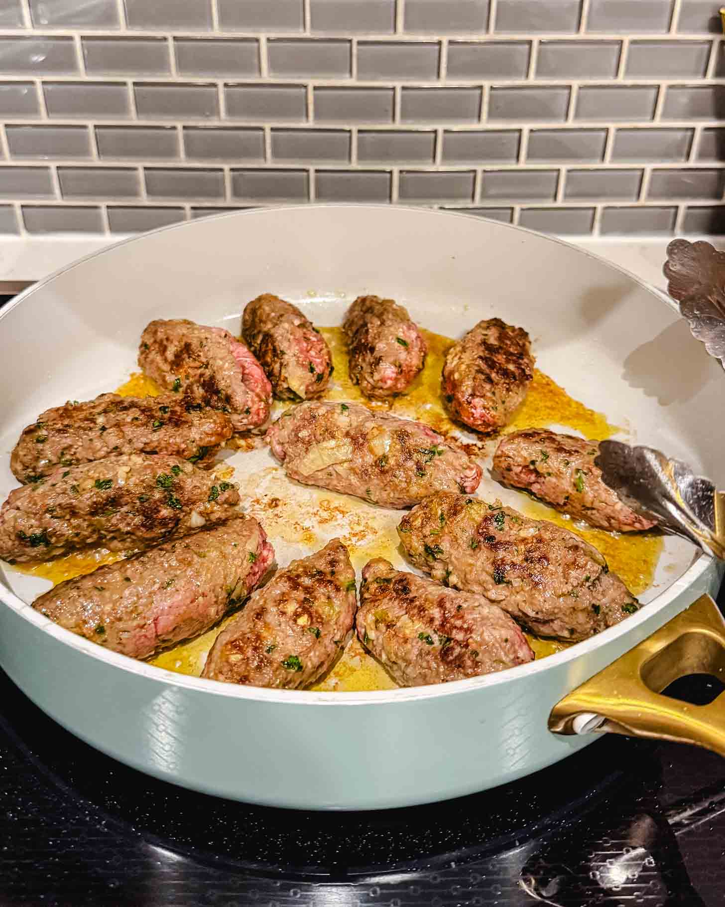 A white pan with golden handles contains several browned, oval-shaped pieces of ground meat cooking on a stovetop, with a gray tile backsplash in the background. A pair of metal tongs rests on the pan’s edge.