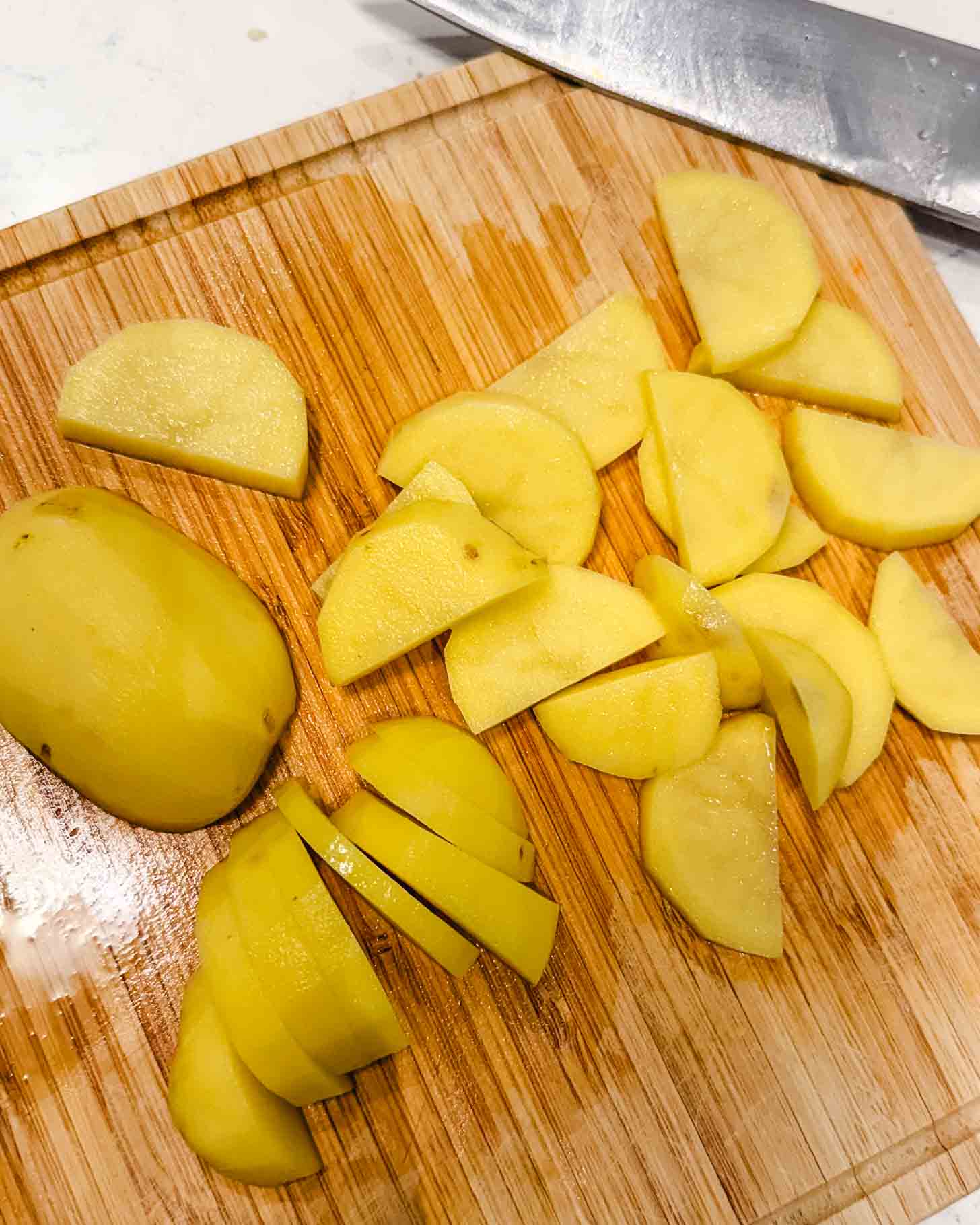 Sliced yellow potatoes on a wooden cutting board with a large knife nearby. Some potato pieces are whole, while others are cut into thin, semi-circular slices.