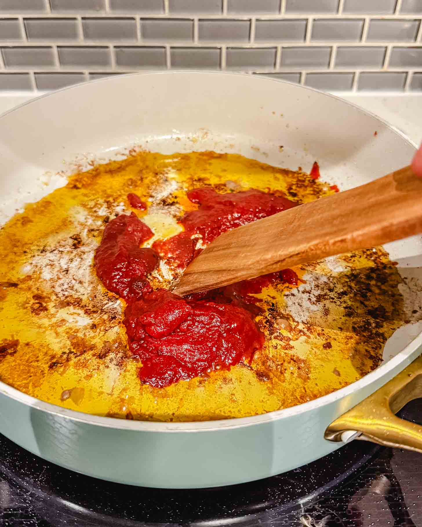 A person uses a wooden spatula to stir tomato paste in a white skillet with oil and browned bits on the stove, with a gray tile backsplash in the background.