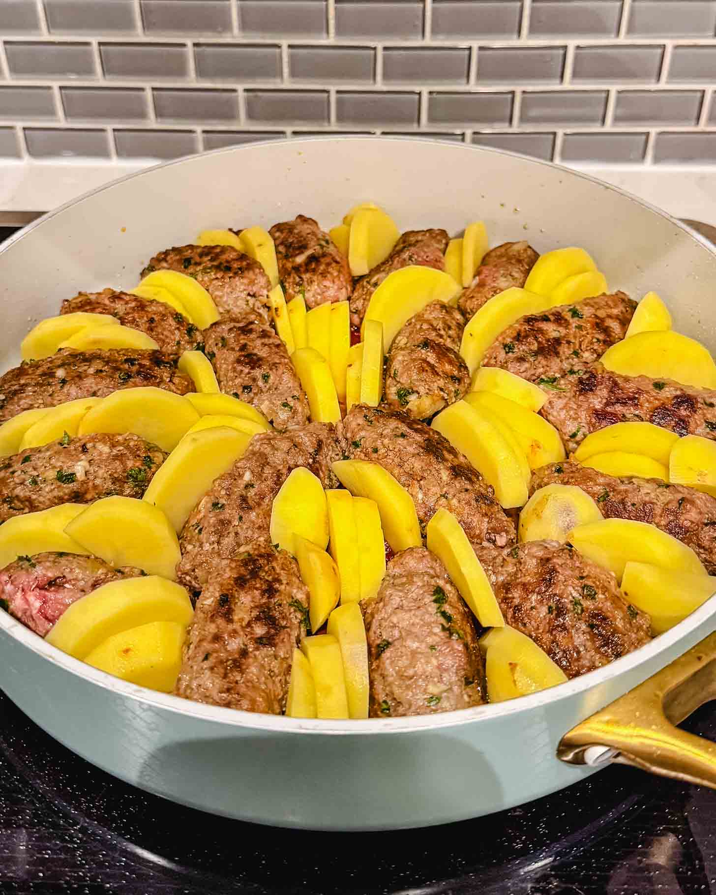A round pan on a stovetop filled with browned oval-shaped meat patties and yellow potato wedges arranged in alternating sections, ready to be cooked, with a gray subway tile backsplash in the background.