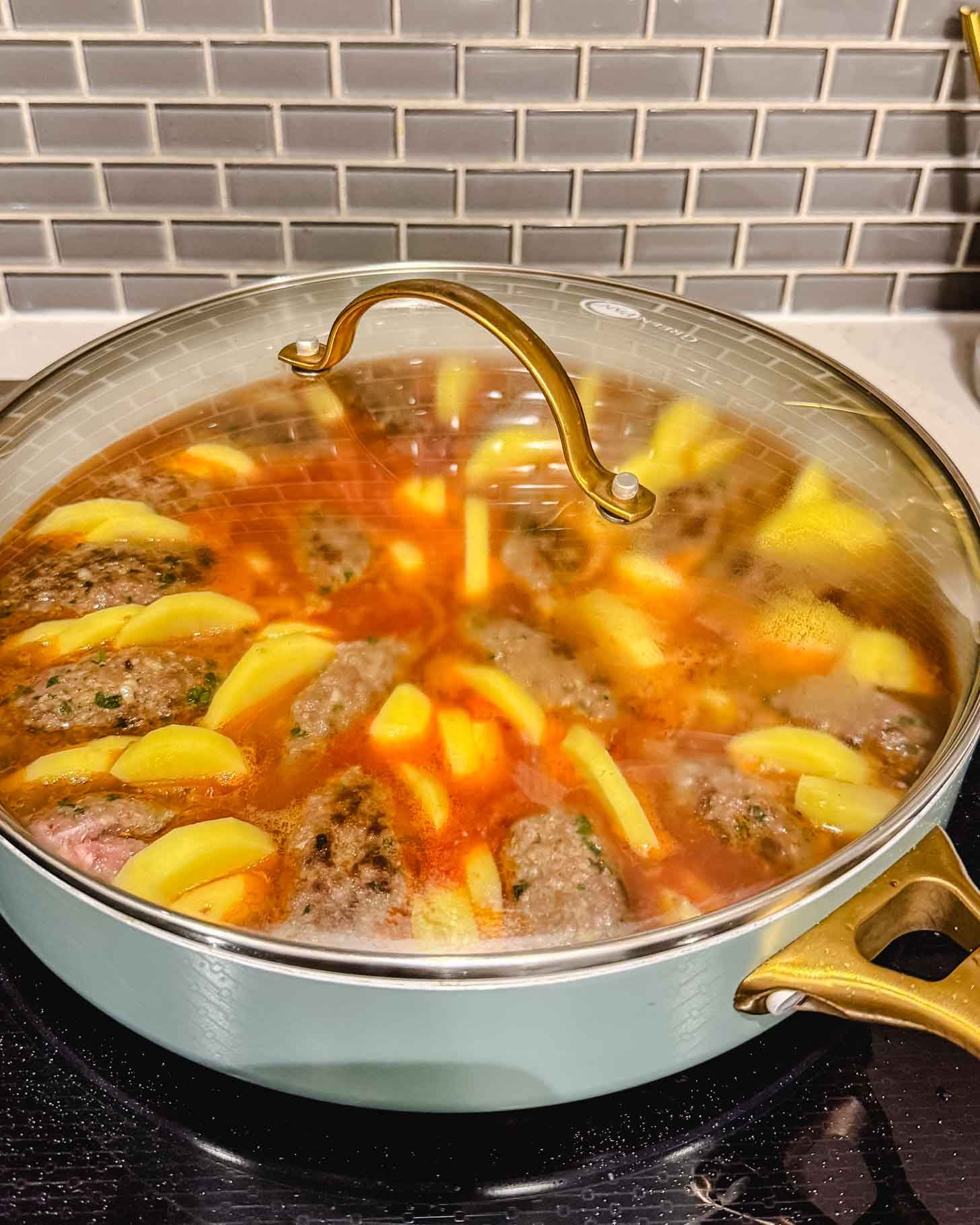 A lidded pan on a stove contains simmering meat patties, sliced potatoes, and carrots in a reddish broth. The background shows a gray tiled backsplash and a white countertop.