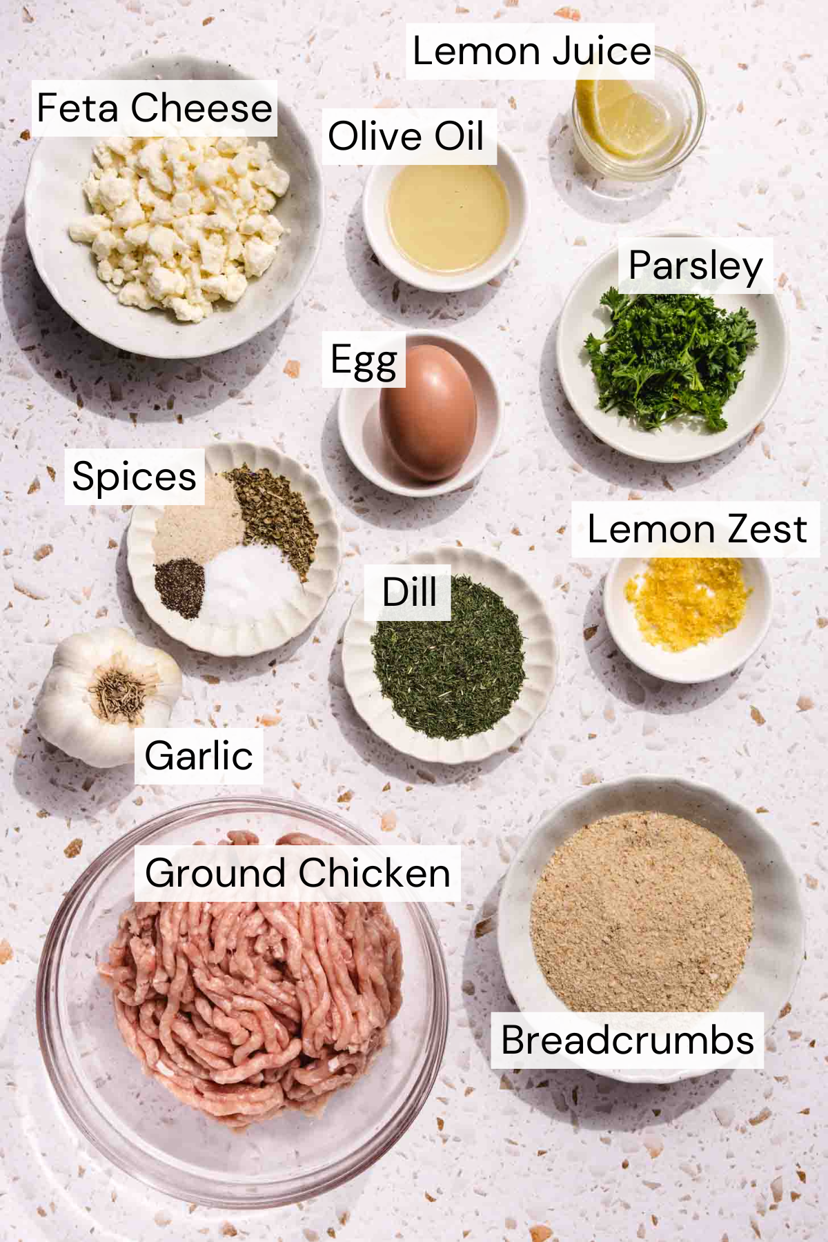 A flat lay of labeled ingredients in bowls for a recipe, including feta cheese, olive oil, lemon juice, parsley, egg, spices, dill, lemon zest, garlic, ground chicken, and breadcrumbs on a light surface.