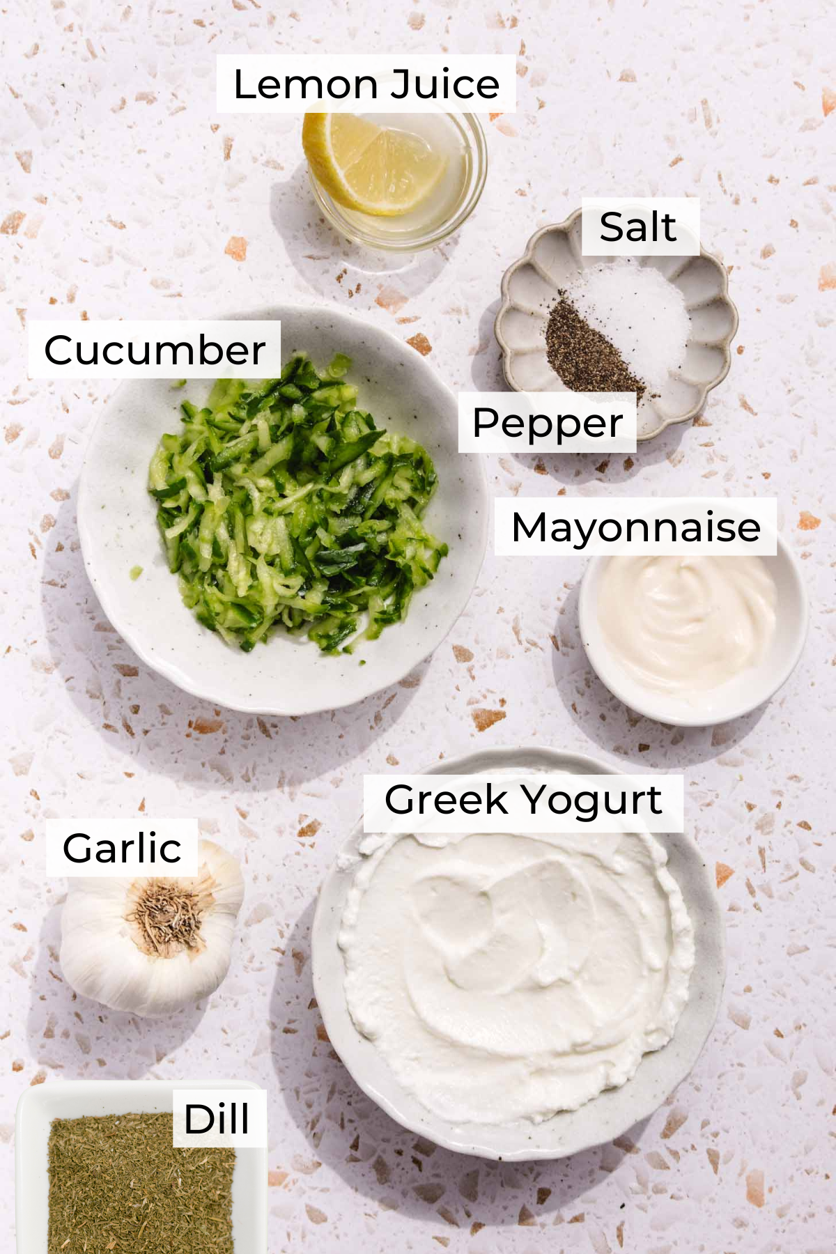 Bowls arranged on a countertop containing labeled ingredients for tzatziki: lemon juice, salt, pepper, grated cucumber, mayonnaise, Greek yogurt, garlic, and dried dill.