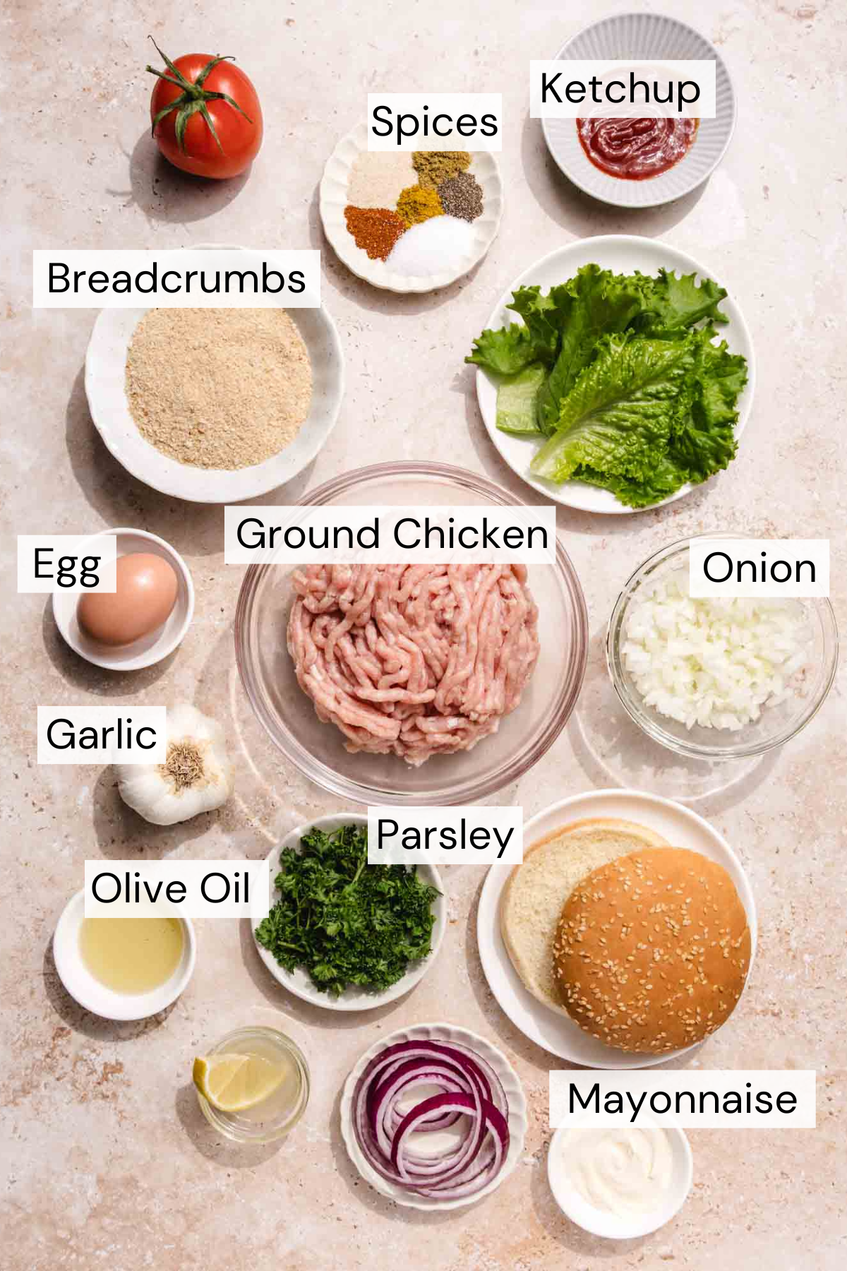 Labeled ingredients for chicken burgers are arranged on a countertop, including ground chicken, lettuce, onion, breadcrumbs, parsley, egg, garlic, olive oil, spices, ketchup, mayonnaise, and a burger bun.