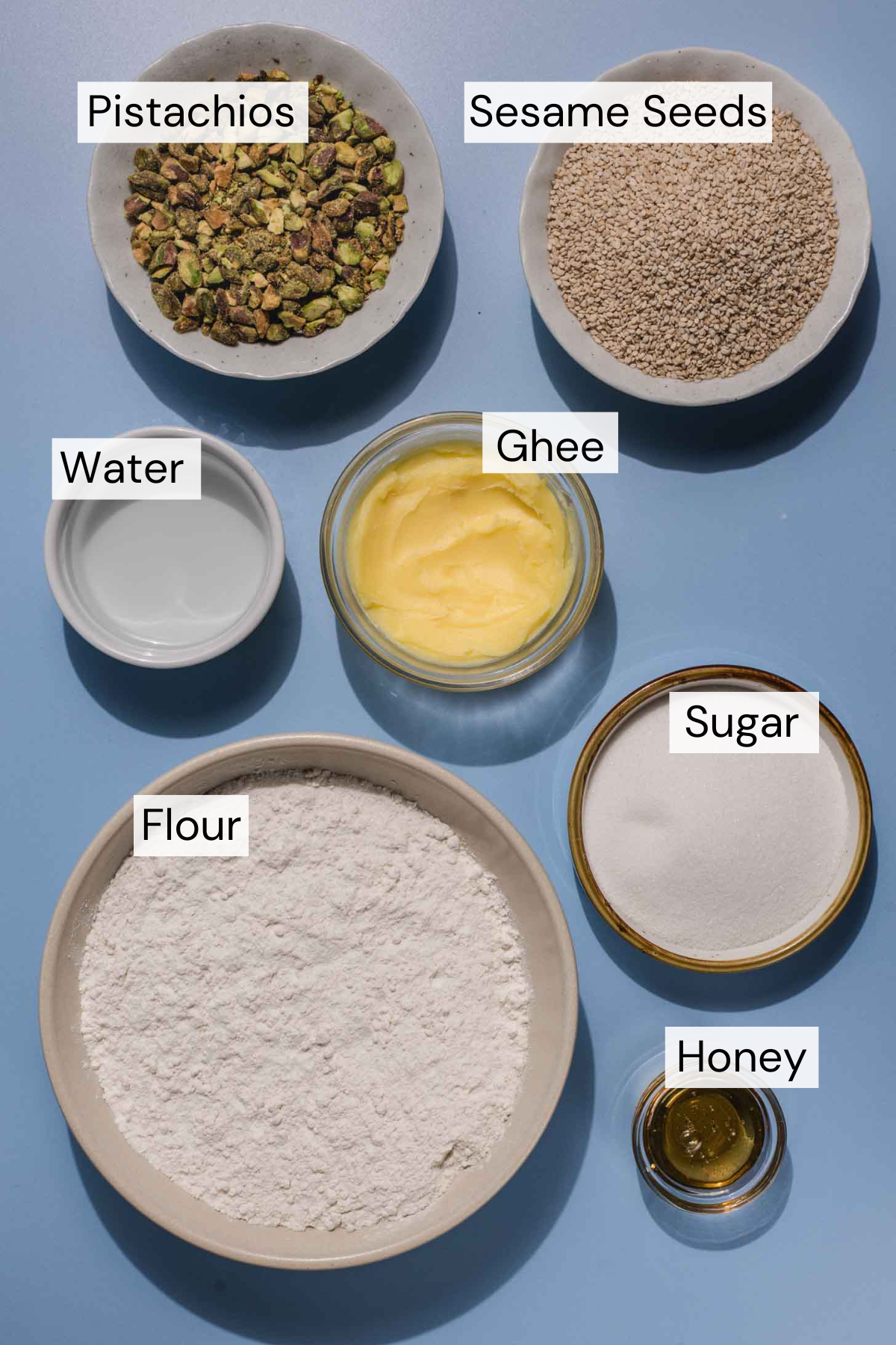 A top-down view of bowls containing labeled ingredients: pistachios, sesame seeds, water, ghee, flour, sugar, and honey, all arranged on a blue surface.