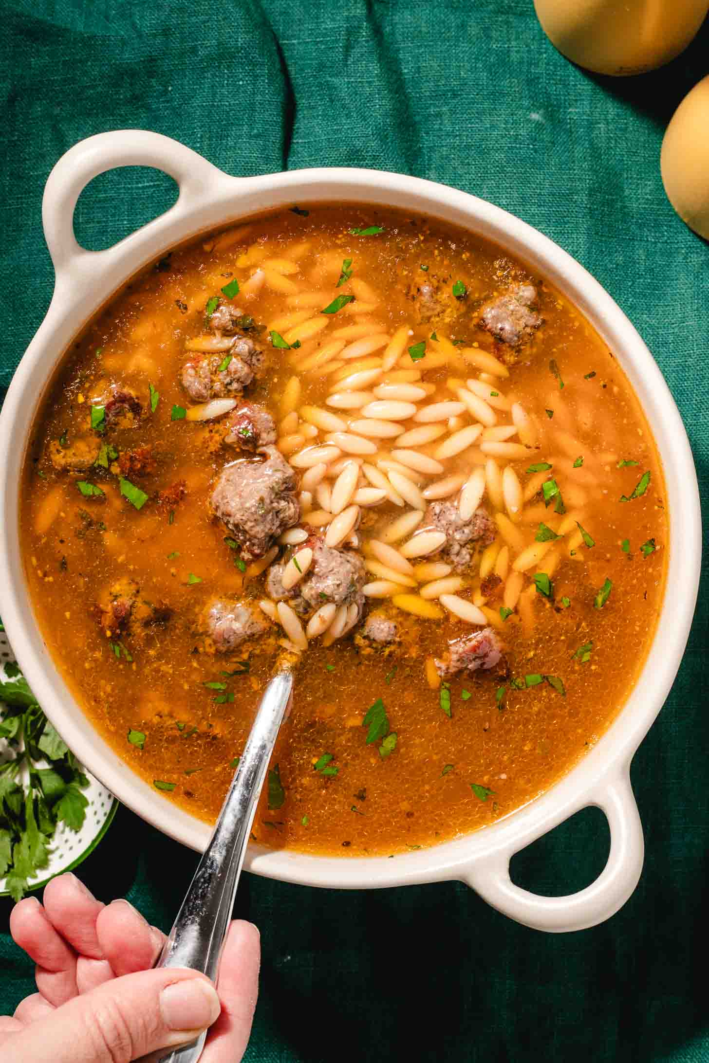 A hand holds a spoon in a white bowl filled with soup containing meatballs, orzo pasta, and chopped herbs, set on a green tablecloth.