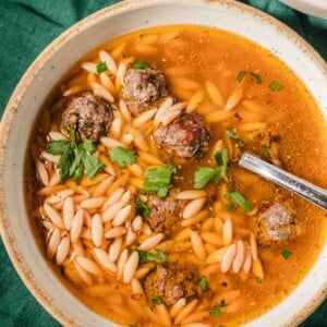 A bowl of soup with small meatballs, orzo pasta, and fresh parsley garnish sits on a green cloth with a spoon in the bowl. The broth is light and clear, showing the pasta and meatballs clearly.
