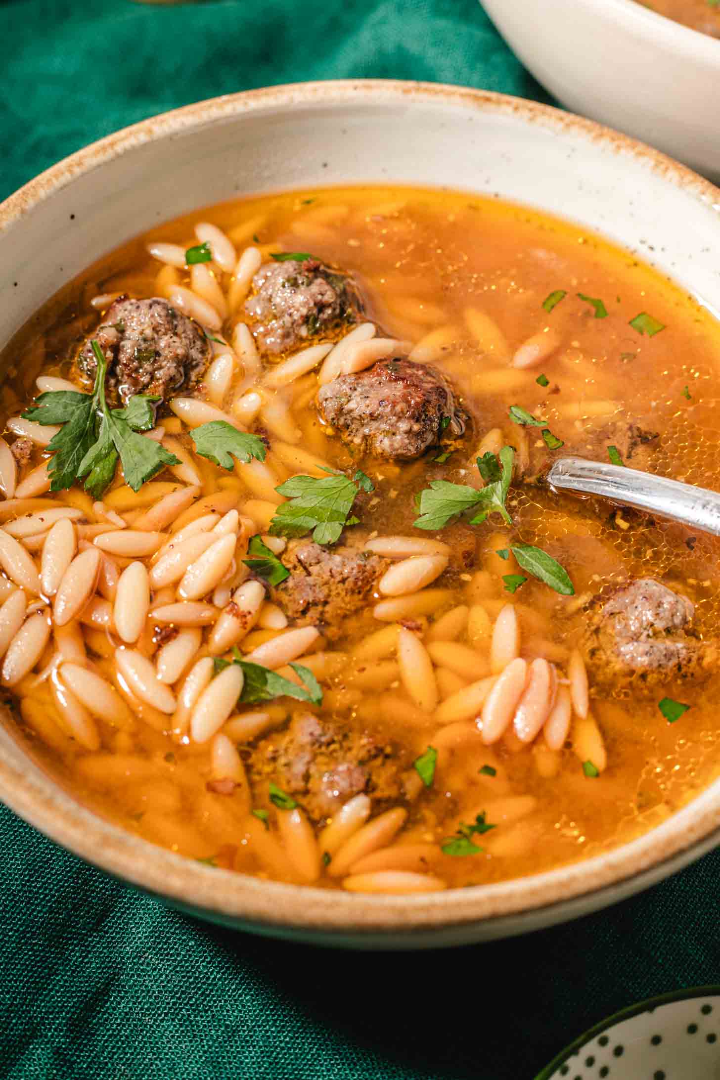 A bowl of soup with meatballs, orzo pasta, and fresh parsley garnish sits on a green cloth. A spoon rests in the bowl, and the broth appears golden and flavorful.
