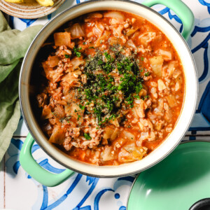 A pot of hearty stew with ground meat, chopped vegetables, and tomato sauce, garnished with fresh herbs, sits on a patterned blue and white surface next to lemon wedges and a green cloth.