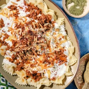 A platter of bowtie pasta topped with seasoned ground meat, creamy white sauce, slivered almonds, herbs, and spices, with small bowls of salt and dried herbs on the side.