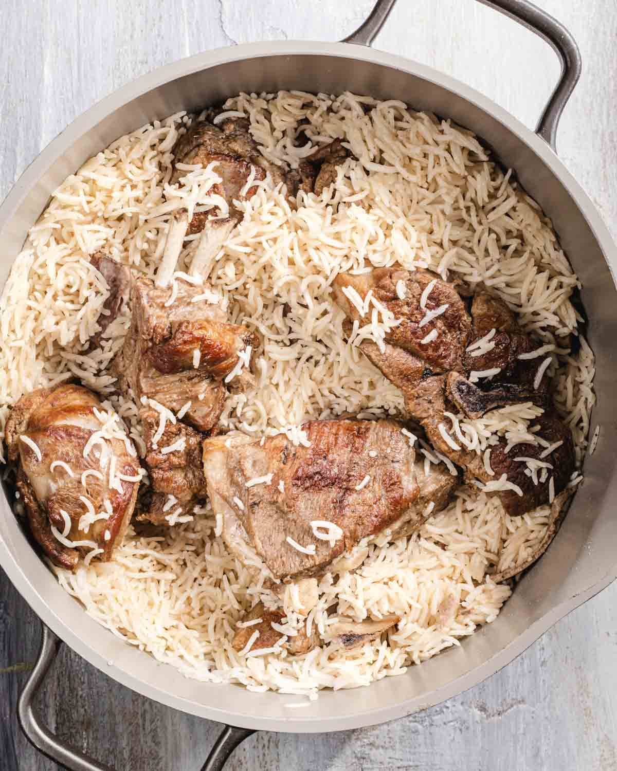 A large metal pot filled with cooked basmati rice and several browned pieces of meat, likely lamb or beef, on a light-colored surface.