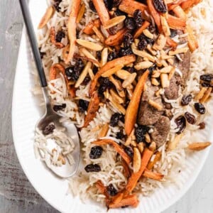 A white oval plate of rice topped with sautéed carrots, slivered almonds, raisins, and pieces of meat. A serving spoon rests on the side of the plate.