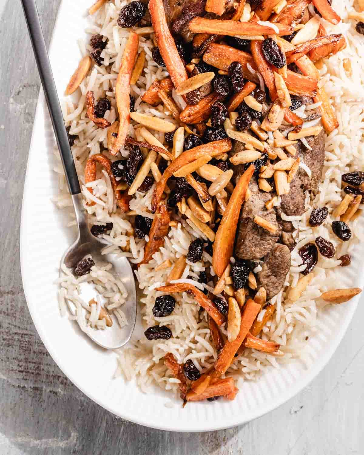 A white oval plate of rice topped with sautéed carrots, slivered almonds, raisins, and pieces of meat. A serving spoon rests on the side of the plate.