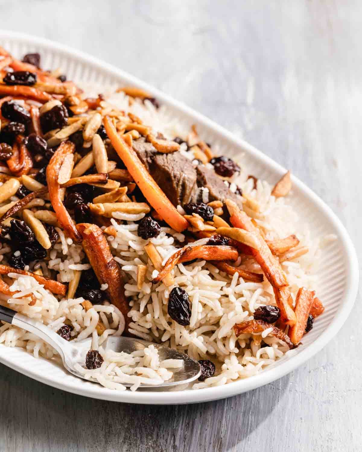 A plate of rice topped with sliced carrots, raisins, slivered almonds, and pieces of meat, with a serving spoon placed on the side.
