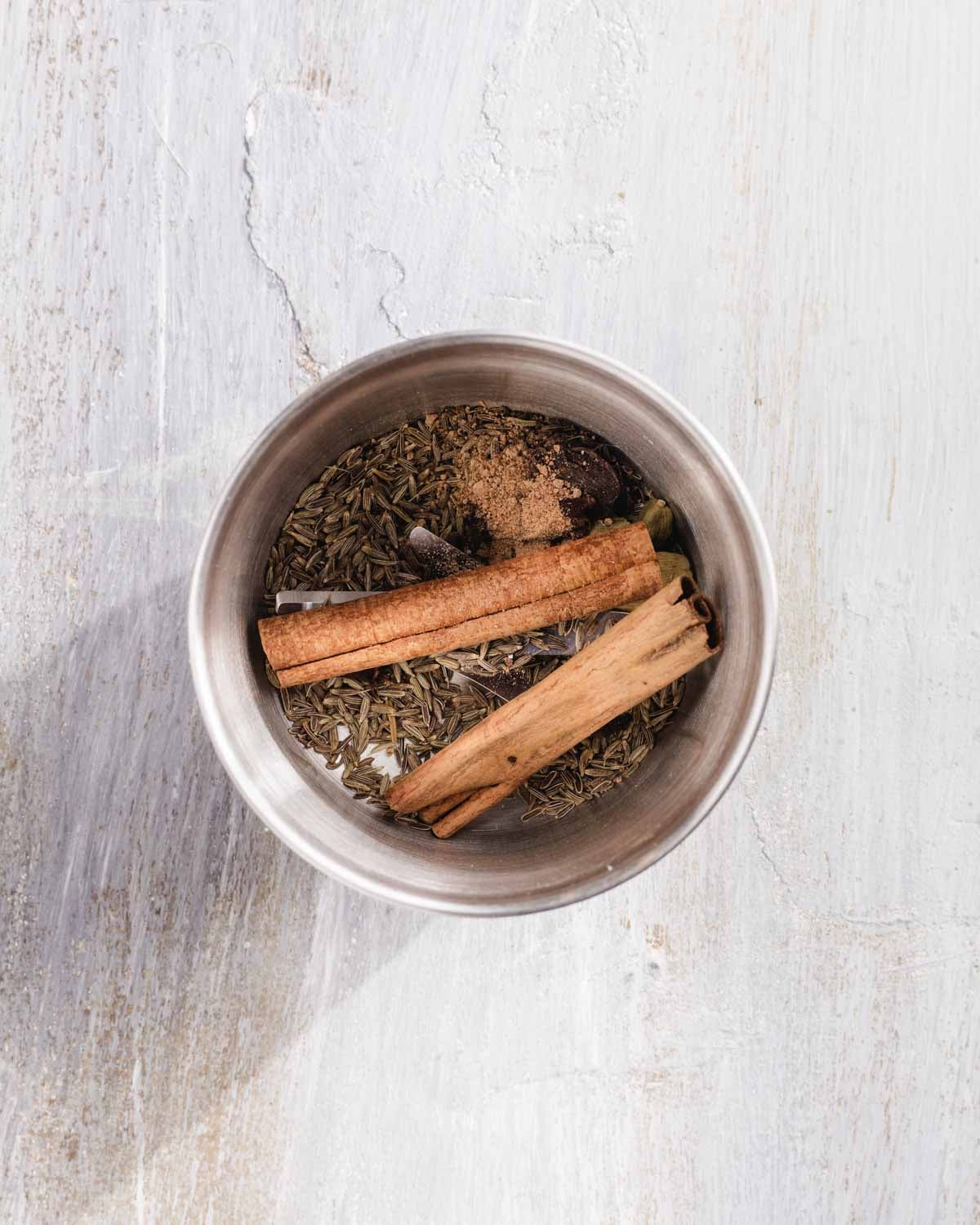 A metal bowl containing whole spices, including cinnamon sticks, cloves, cardamom pods, cumin seeds, and a small mound of ground spice, sits on a light wooden surface.