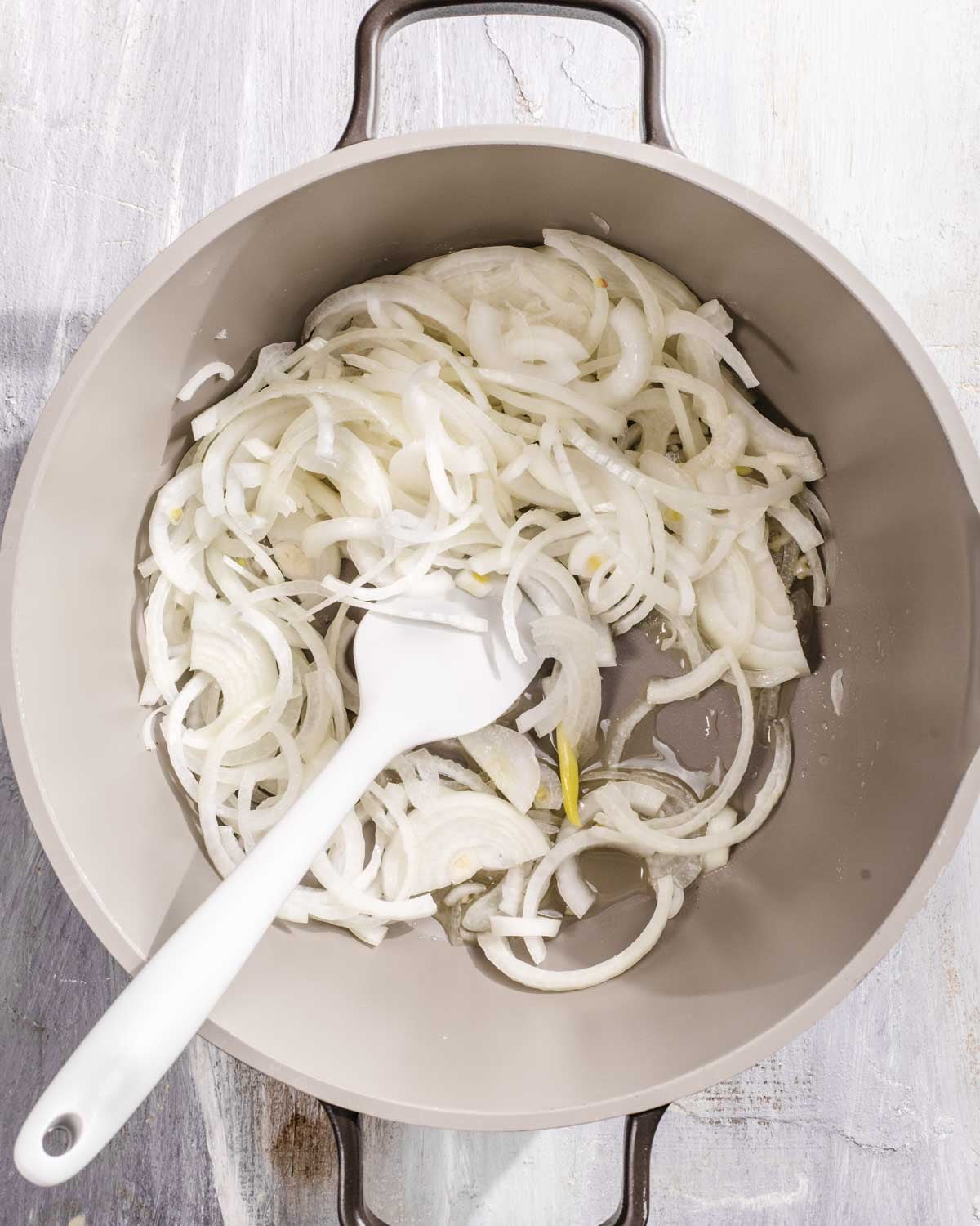 Thinly sliced onions being stirred with a white spatula in a light-colored pan with two handles, sitting on a pale wooden surface.