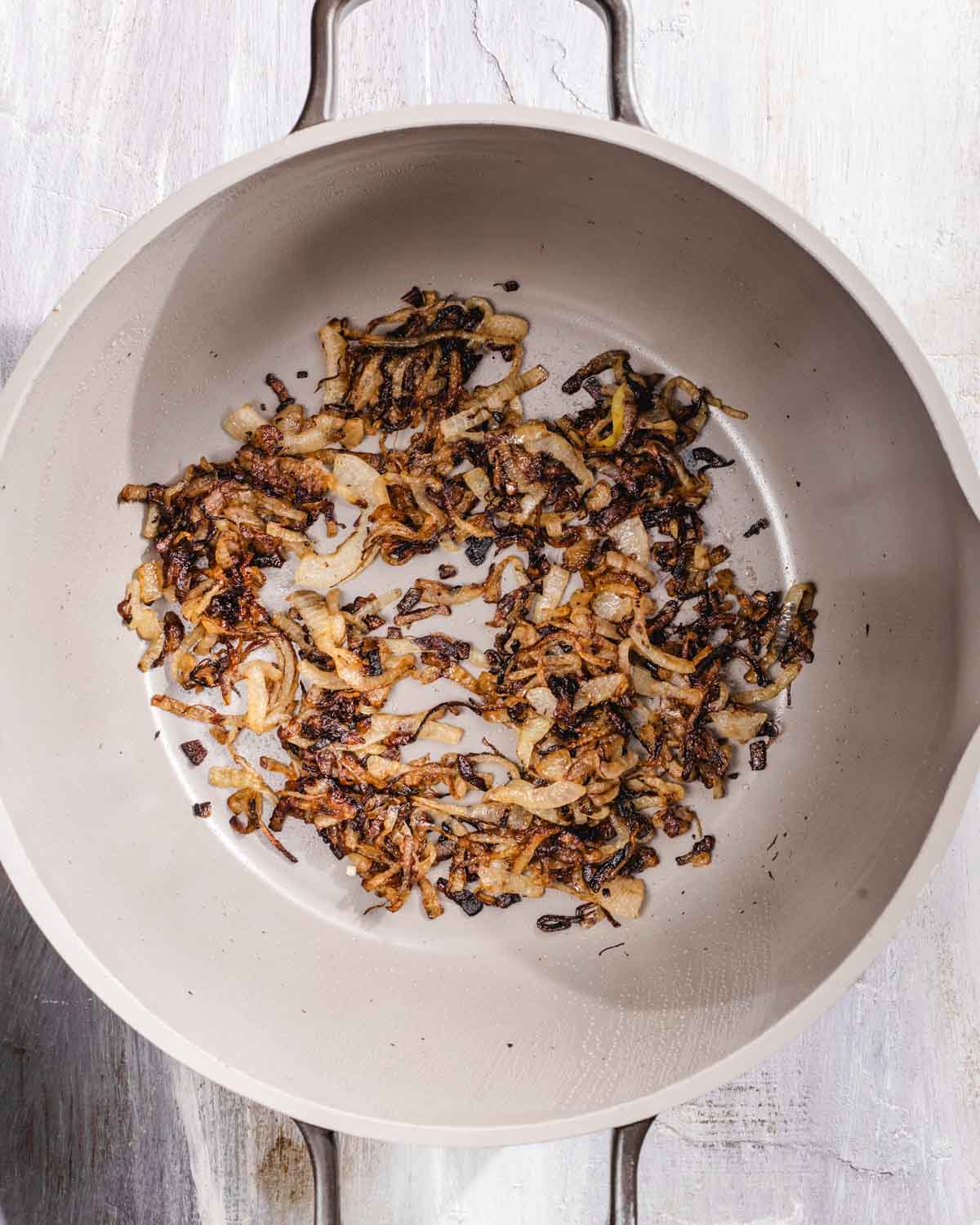 Sliced onions being caramelized in a light-colored, round pot on a white surface, with some pieces browned and others golden.