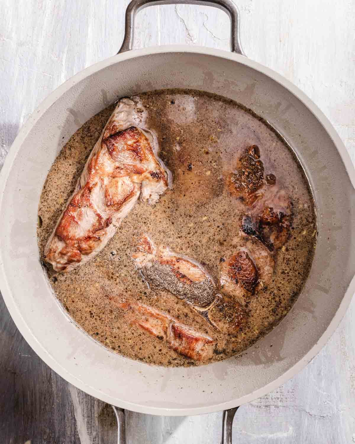 A white pot filled with browned pork pieces simmering in a light brown broth on a light-colored surface.