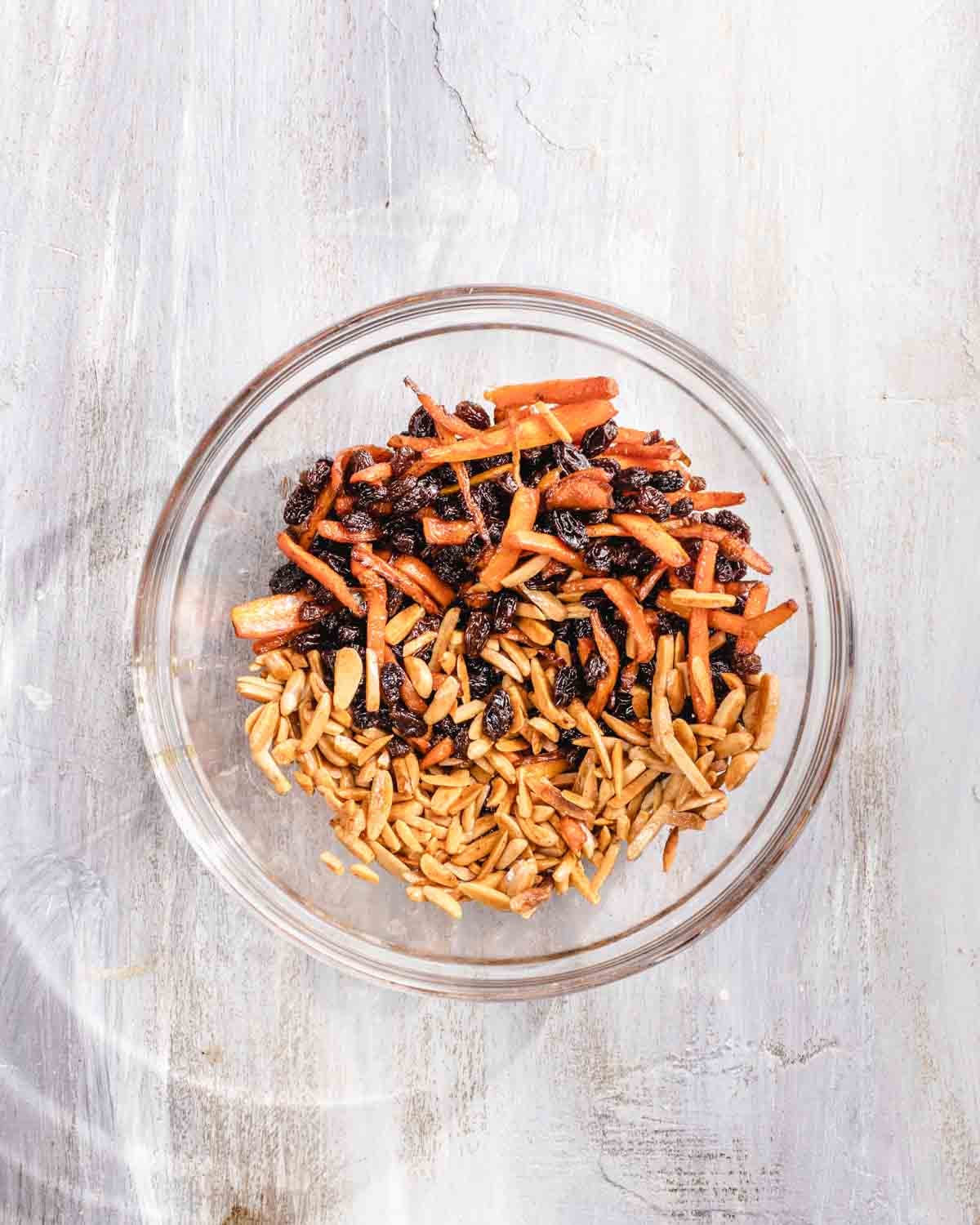A glass bowl containing a mixture of slivered almonds, shredded sautéed carrots, and black raisins, placed on a light wooden surface.