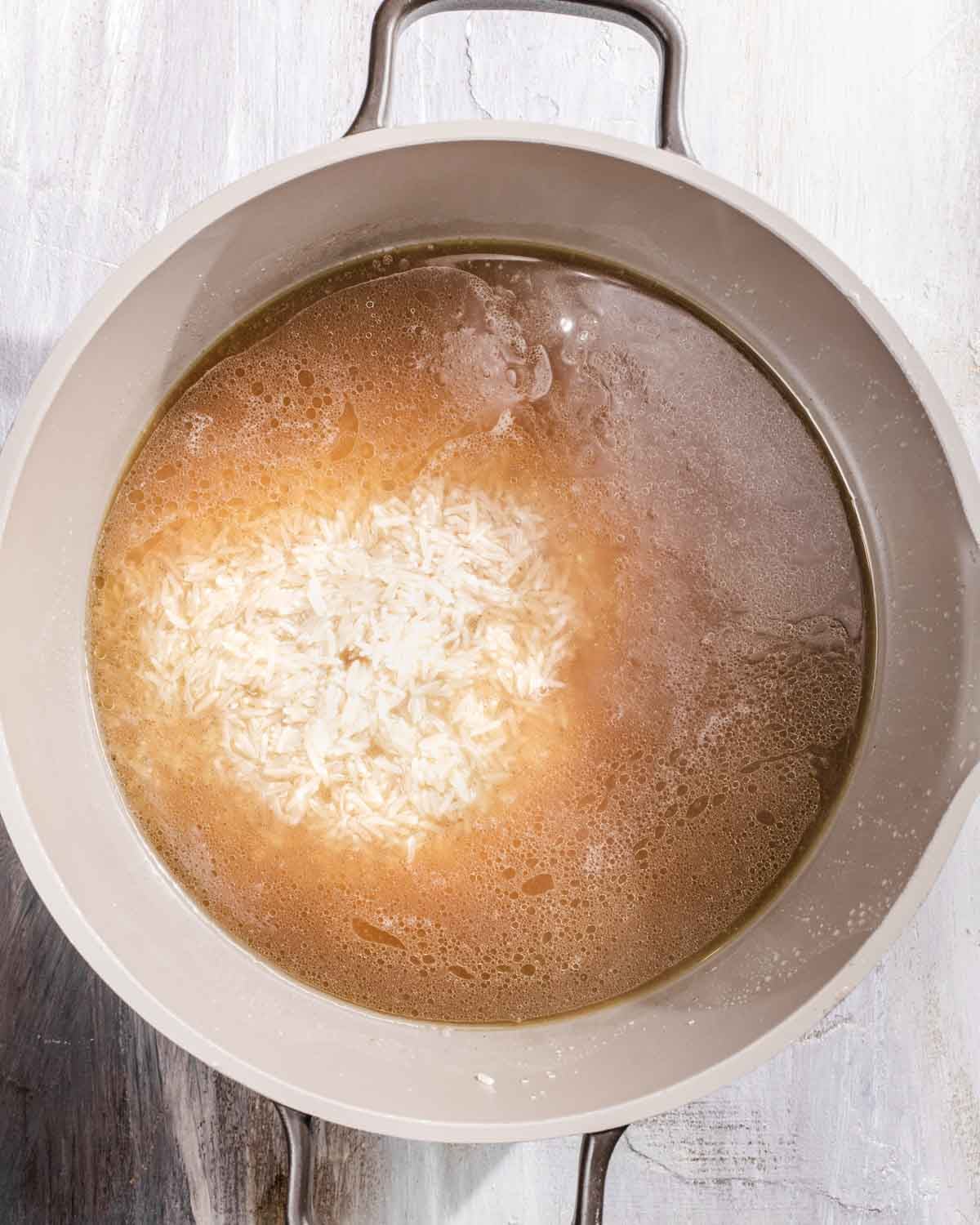 A pot filled with water, uncooked white rice, and broth is placed on a light wooden surface, ready for cooking.
